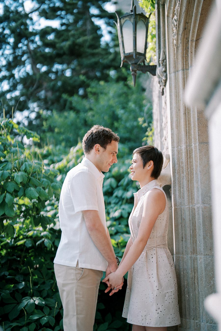 A couple holding hands and smiling at each other outdoors, surrounded by greenery and standing by a stone wall with an ornate lamp.