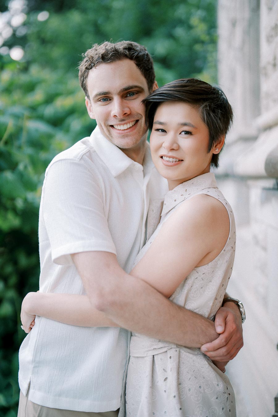 A happy couple embracing outdoors, both smiling and wearing light-colored clothing, surrounded by greenery and stone architecture.