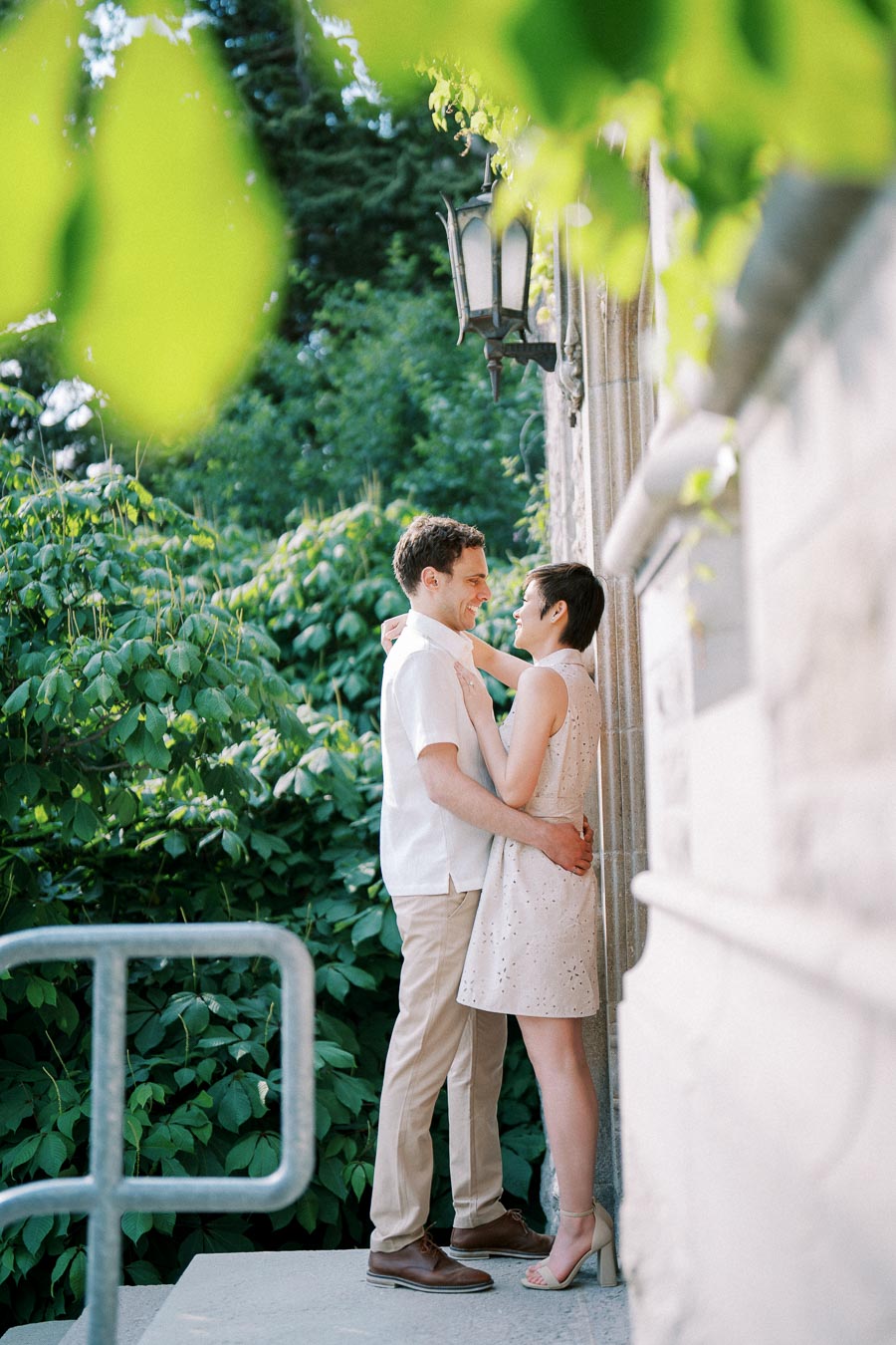 Couple embracing outdoors in a lush garden setting, standing close to a stone building surrounded by greenery and soft natural light.