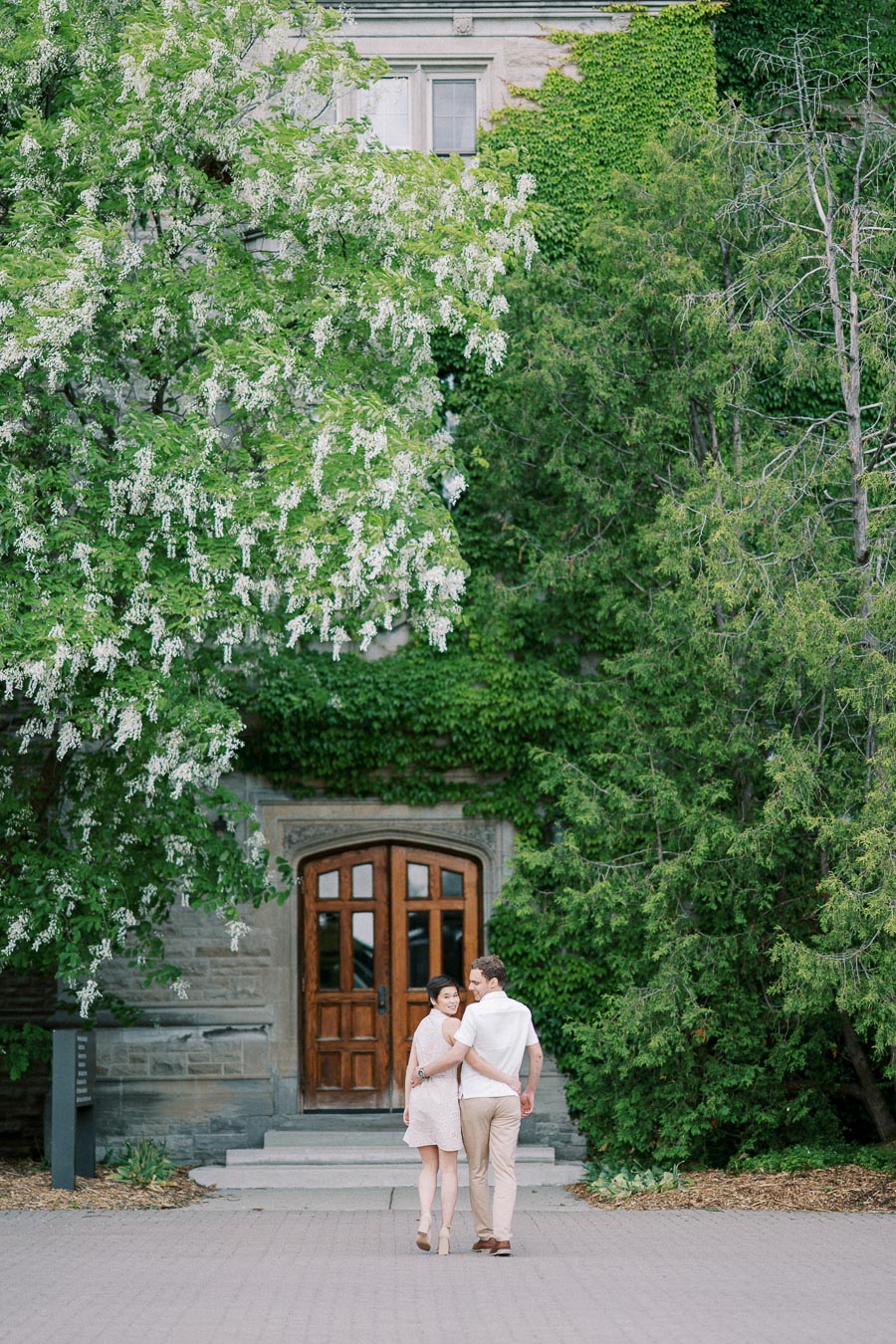 A couple walking arm in arm towards a historic stone building with a wooden door, surrounded by lush green trees and ivy-covered walls.