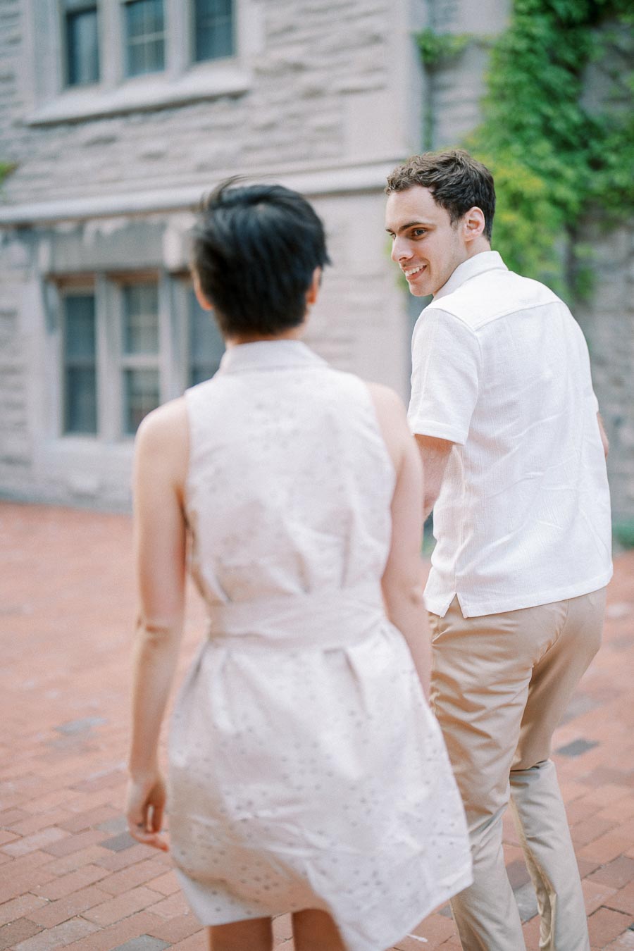 A couple smiling while walking on a brick pathway near a historic stone building covered in ivy.