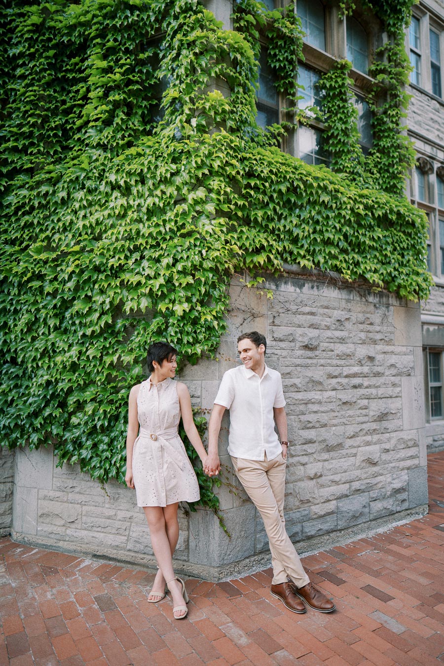 A couple holding hands and smiling in front of an ivy-covered stone building.