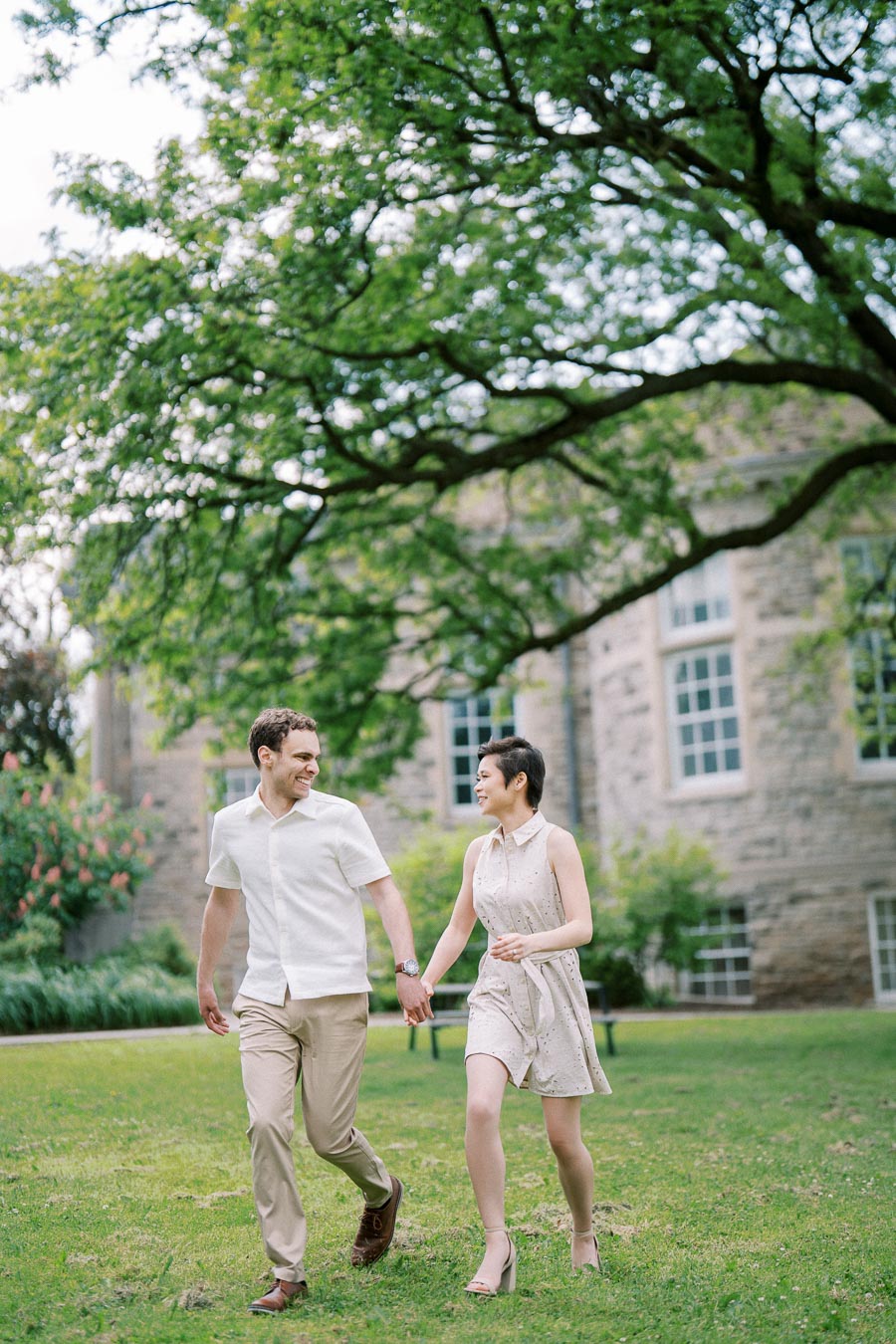 A happy couple holding hands and walking on a lush green lawn, with a quaint stone building and vibrant trees in the background, capturing a serene and joyful moment outdoors.