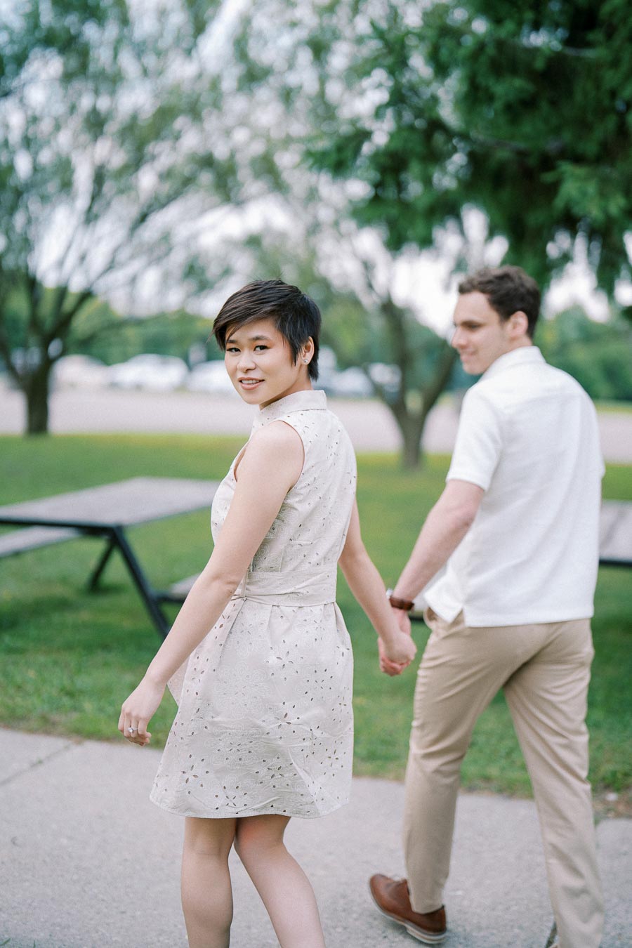 A couple holding hands and walking outdoors in a park, with greenery and trees in the background, wearing light summer clothing and smiling.