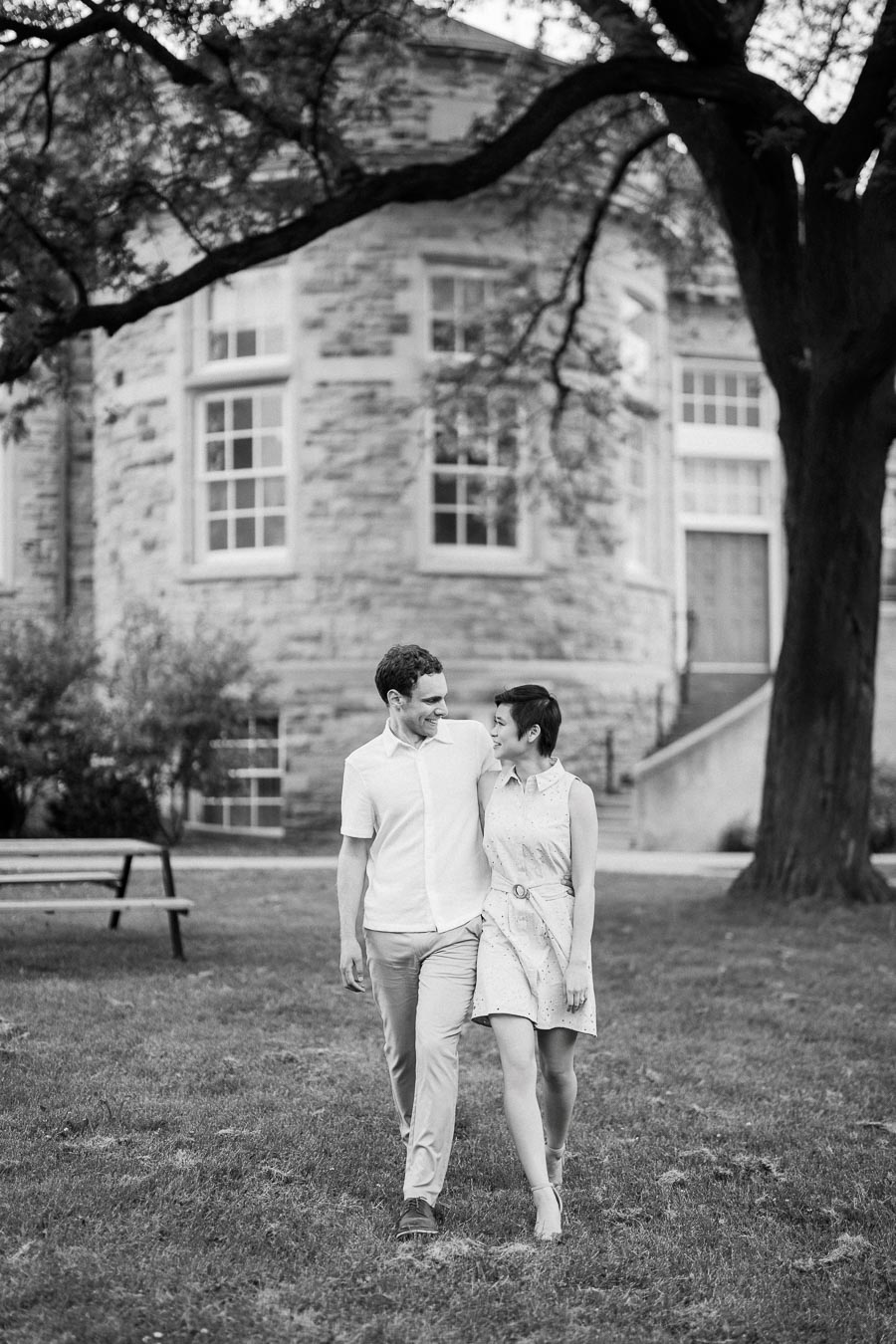 A couple walking arm in arm on a grassy lawn in front of a historic stone building, with large trees casting shade around them, black and white image.