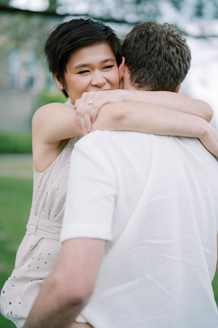 A woman with short hair happily embraces a man in a park, showcasing a sense of joyful affection. She wears a light, sleeveless dress with eyelet patterns, while he is in a white shirt. The background features blurred greenery, adding to the serene atmosphere.