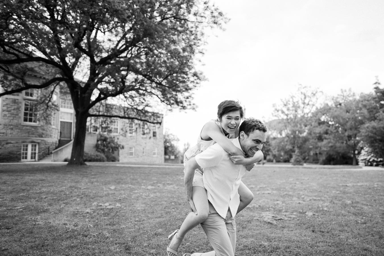 A black and white photo of a joyful couple, with a woman playfully giving a man a piggyback ride in an outdoor setting with grass and trees, and a stone building in the background.