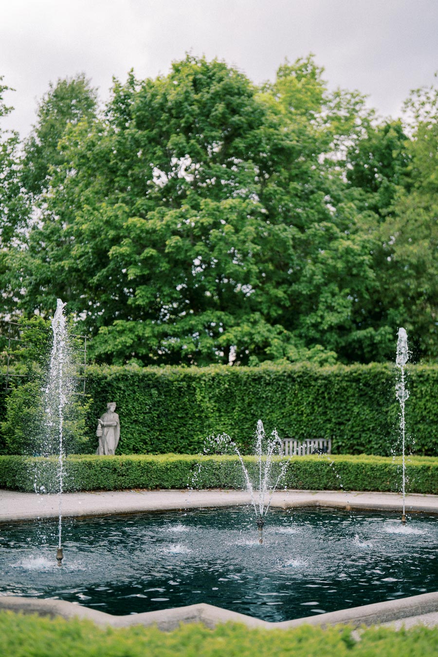 A serene garden scene featuring a round fountain with water jets surrounded by lush greenery and a statue in the background, creating a peaceful and picturesque outdoor environment.