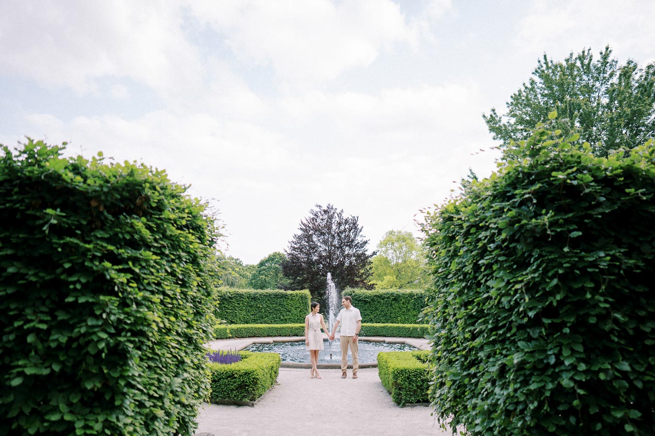 A couple holding hands in a lush garden setting with a central fountain and neatly trimmed hedges, under a partly cloudy sky.