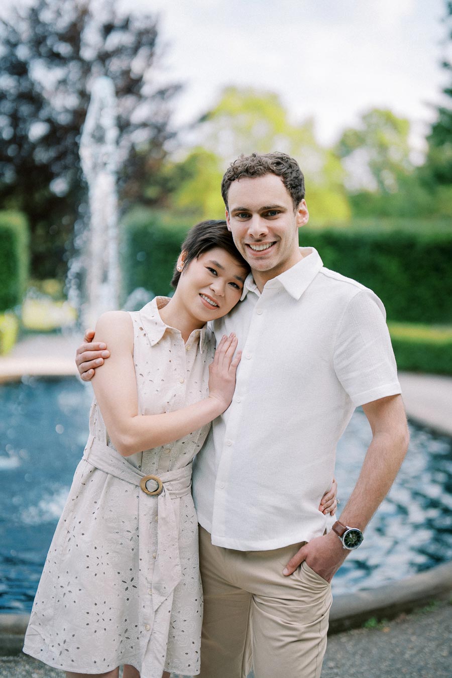 A happy couple embracing in front of a fountain, enjoying a sunny day at a park. Both are smiling warmly, with greenery and bright sky in the background.