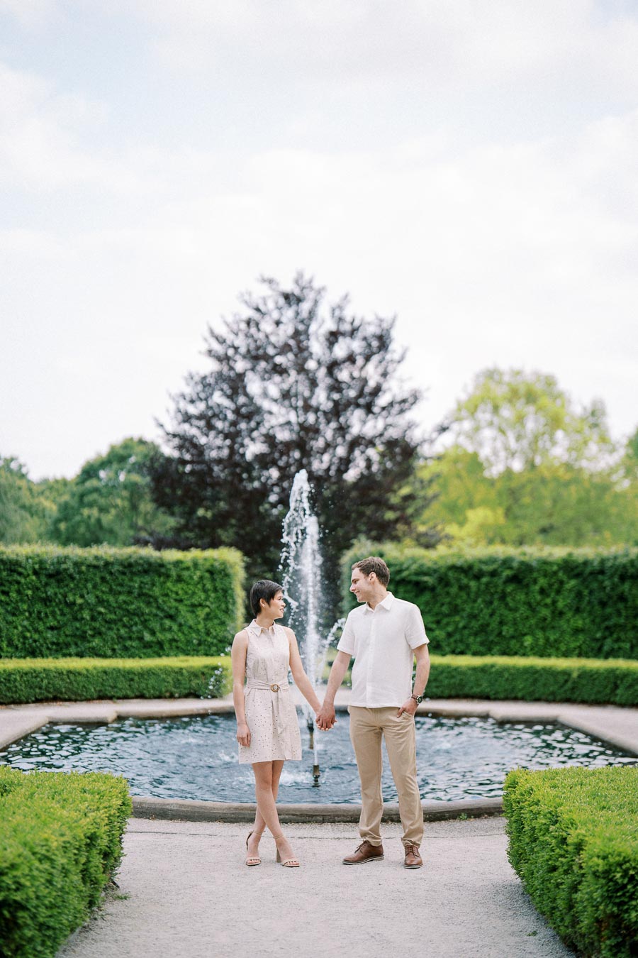 A couple holding hands in front of a fountain in a manicured garden, surrounded by lush green hedges and trees.