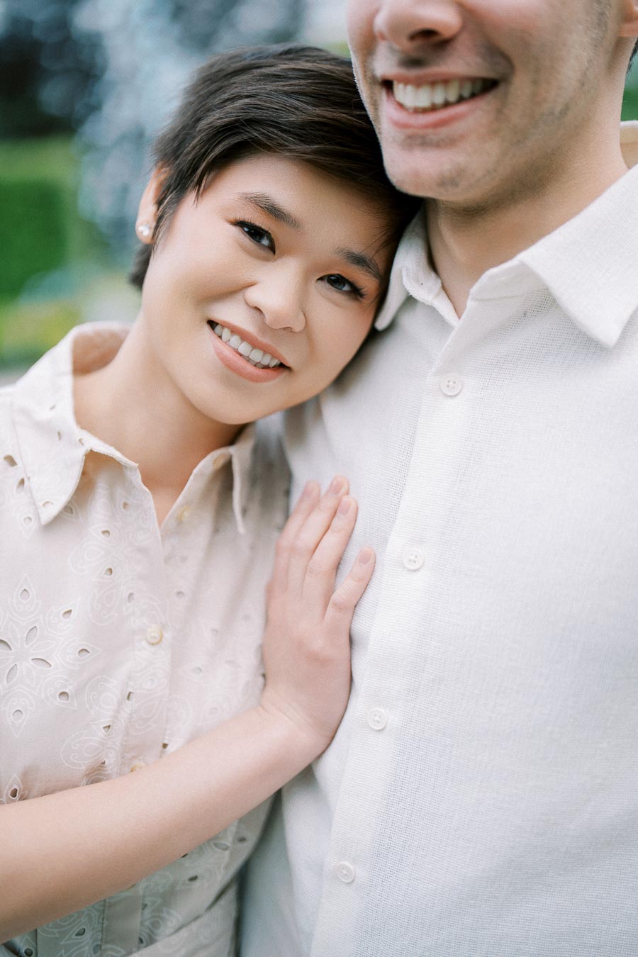 Couple smiling and embracing, woman leaning her head on man’s shoulder, both wearing white shirts, outdoor setting.