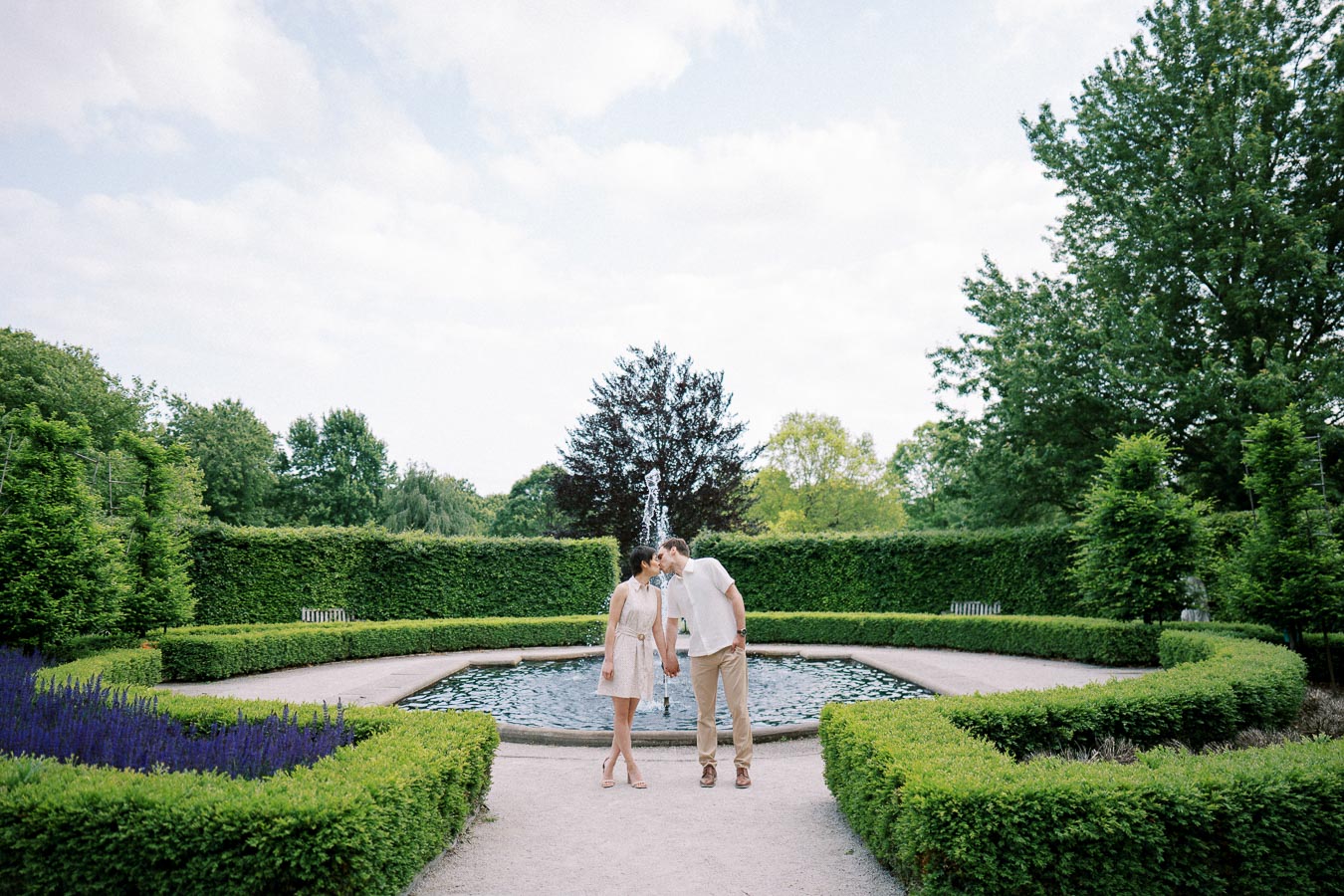 A couple shares a romantic moment while standing near a fountain in a lush green garden, surrounded by manicured hedges and vibrant plants.