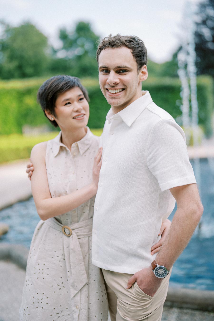 Happy couple posing by a fountain in a garden, both wearing light-colored summer outfits, with greenery in the background.