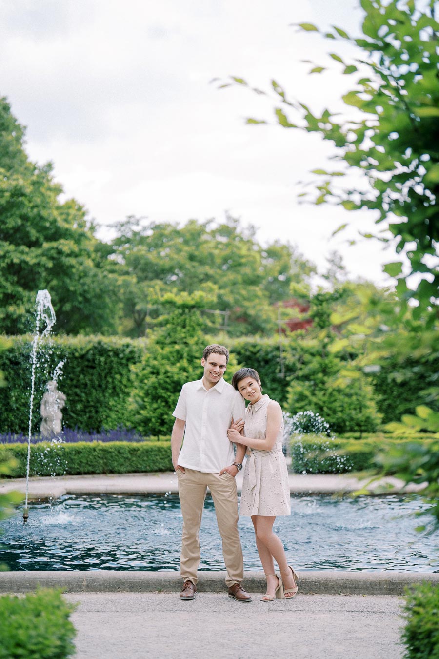 A smiling couple stands together by a fountain in a lush garden, surrounded by greenery and vibrant plants, under a cloudy sky.