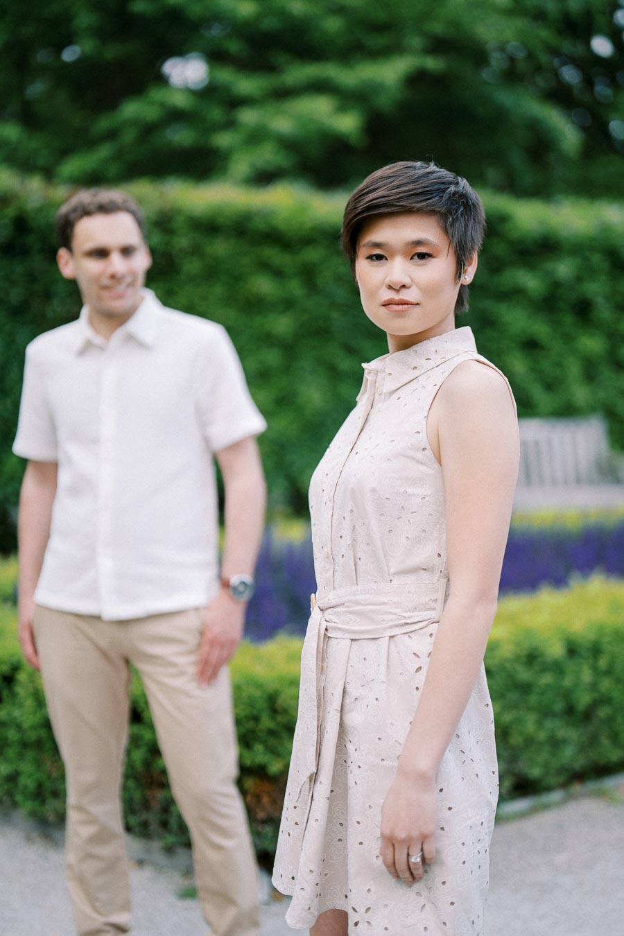 A woman in a patterned sleeveless dress stands in focus in a lush garden while a man in a light-colored shirt and pants is blurred in the background, surrounded by greenery and colorful flowers.