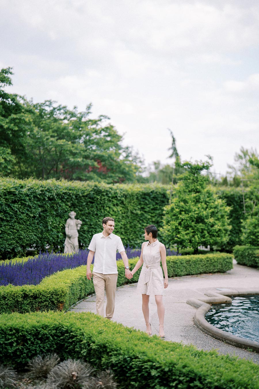 Couple holding hands and walking in a beautifully manicured garden, surrounded by lush greenery and colorful flowers, with a clear blue sky overhead.