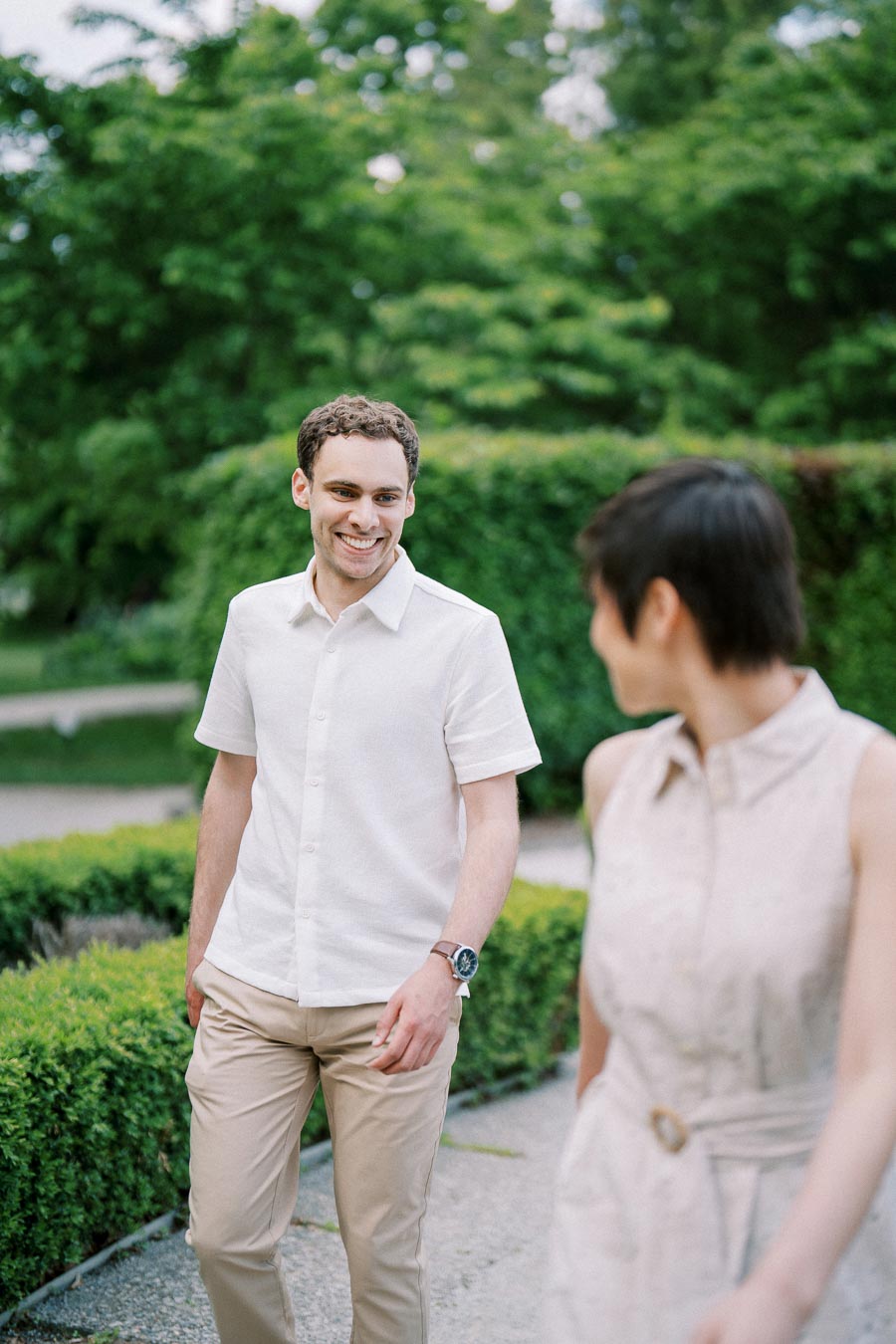 Young man in a white shirt smiling at a woman in a sleeveless dress as they walk through a lush green garden.