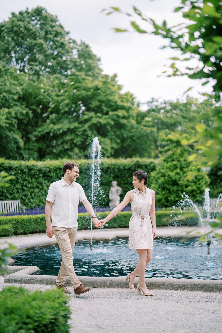 A couple holding hands and walking near a fountain in a lush green garden.
