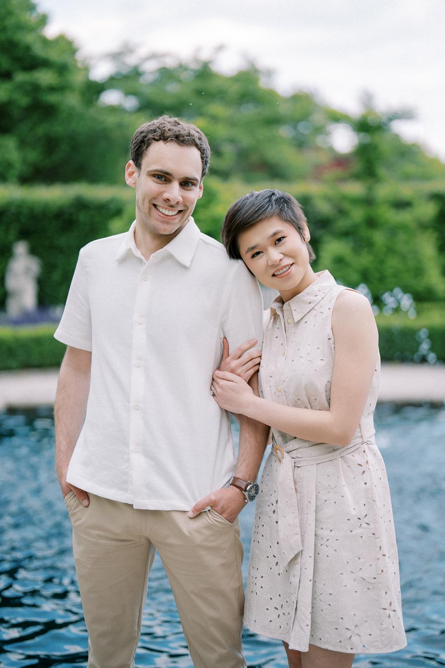 A happy couple posing by a serene pool with lush greenery in the background, showcasing a joyful outdoor moment.