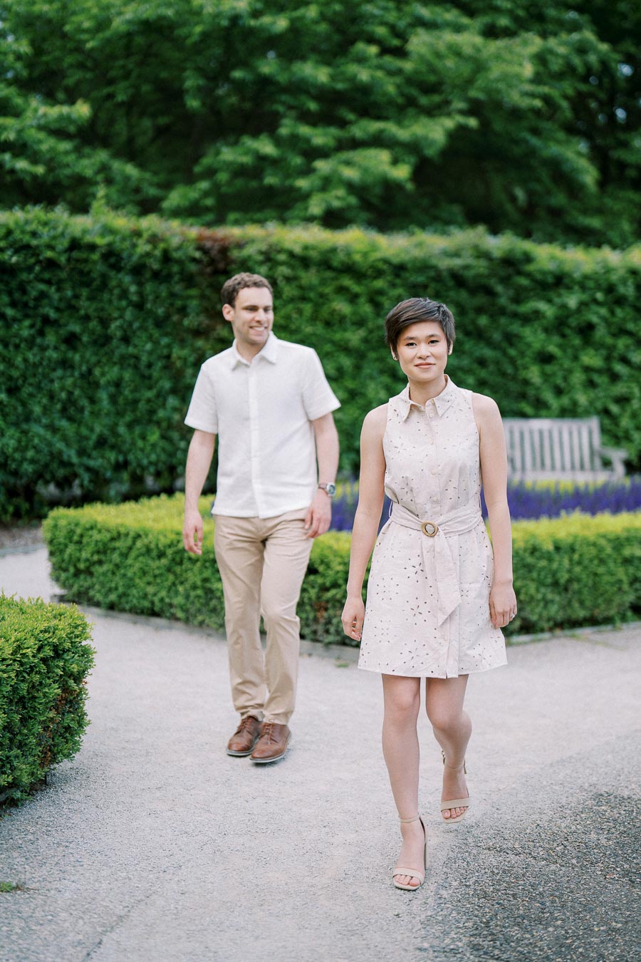 Two people walking in a well-manicured garden, with greenery in the background and a wooden bench partially visible. The woman in a light dress is leading, while the man follows behind in a white shirt and beige pants, both appearing content and relaxed.