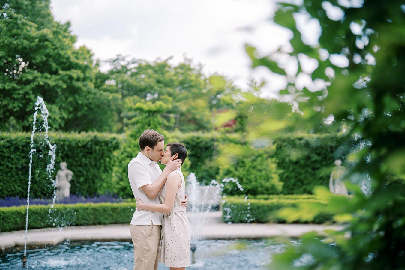 A couple kissing in a lush garden setting beside a fountain, surrounded by green foliage and stone statues, on a bright day.
