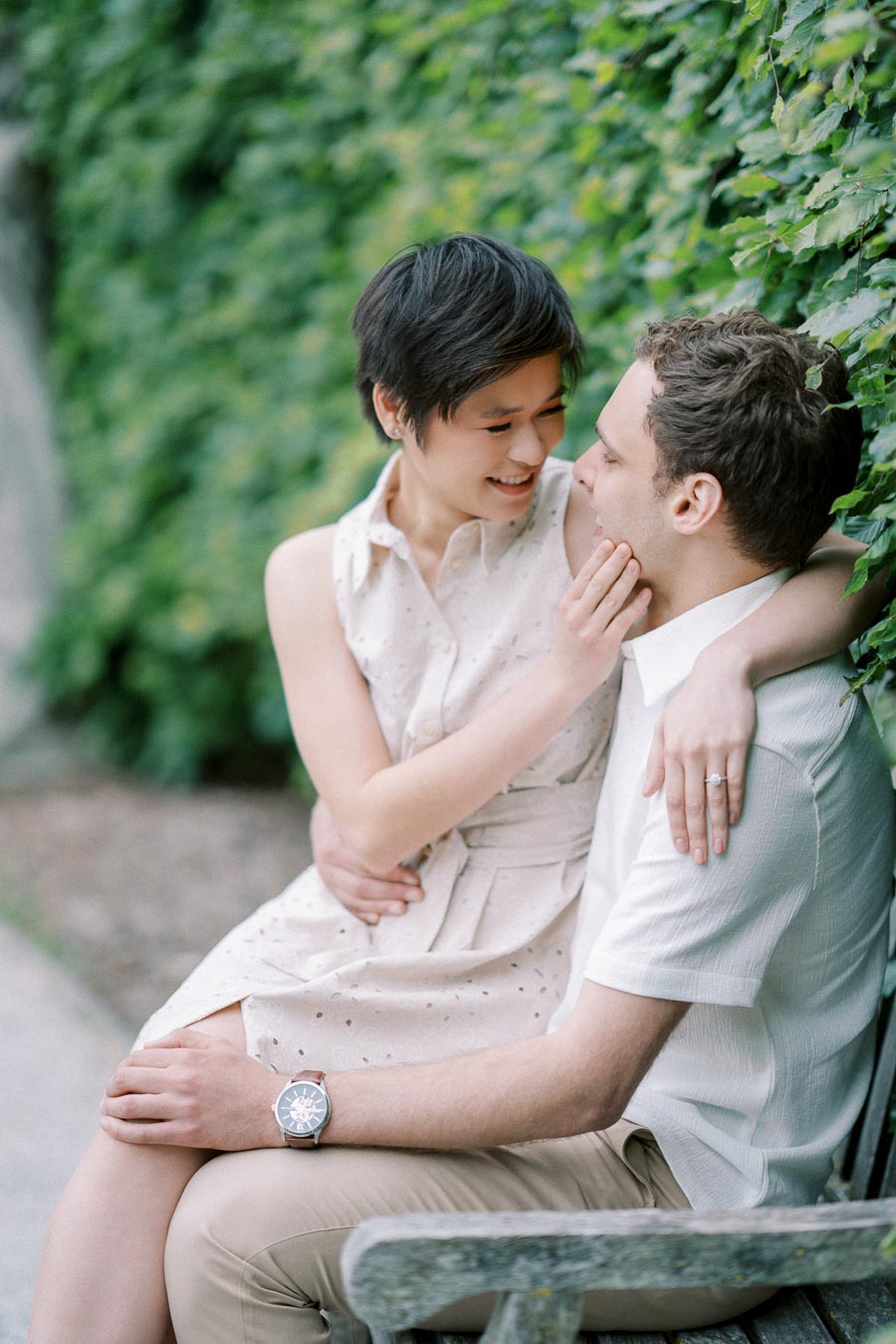 A happy couple sitting on a bench, gazing at each other, surrounded by lush green ivy. The woman is wearing a light dress and has her arm around the man's shoulder, while he is dressed in a white shirt and beige pants.