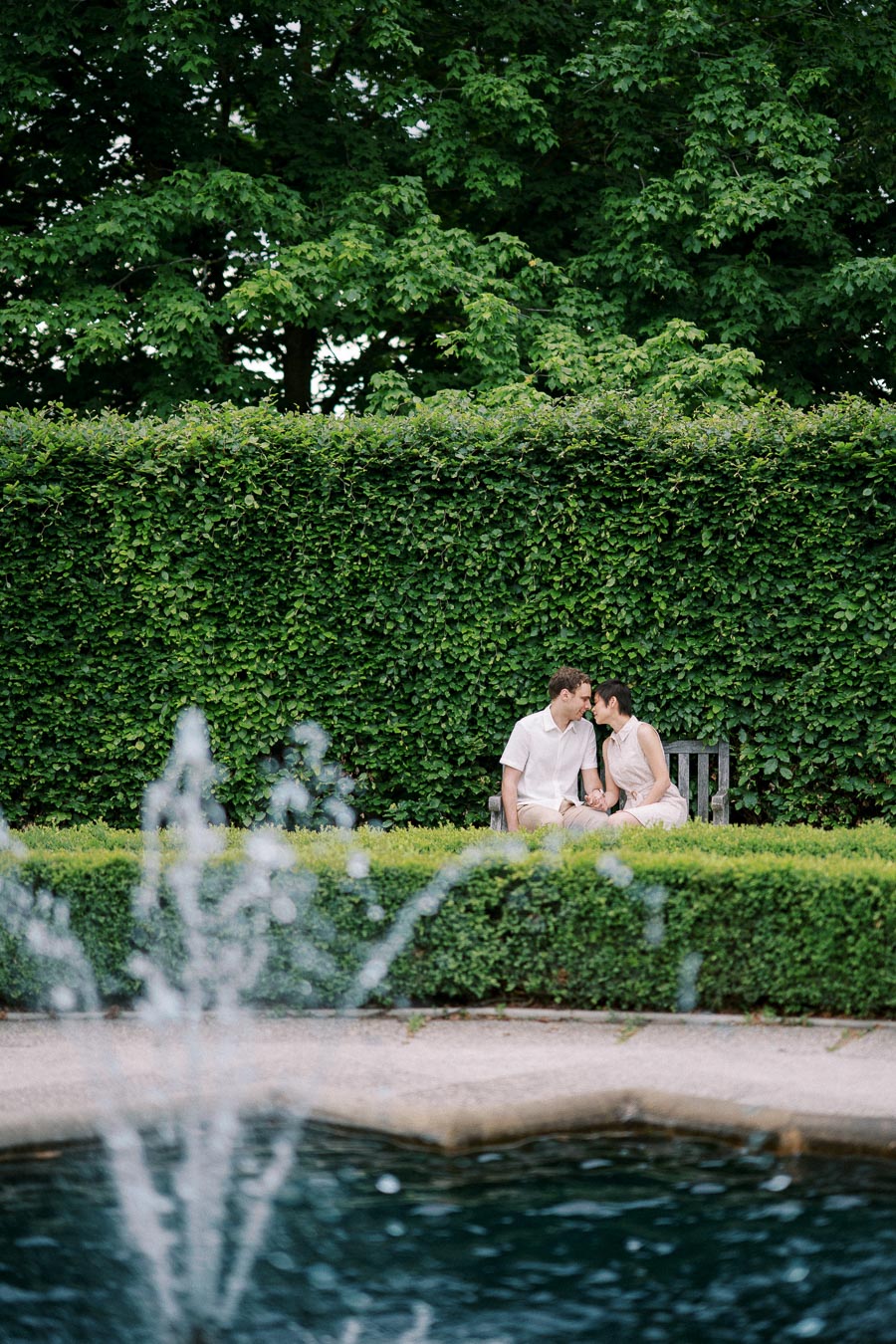 A couple sharing a tender moment on a park bench, surrounded by lush greenery with a fountain in the foreground.