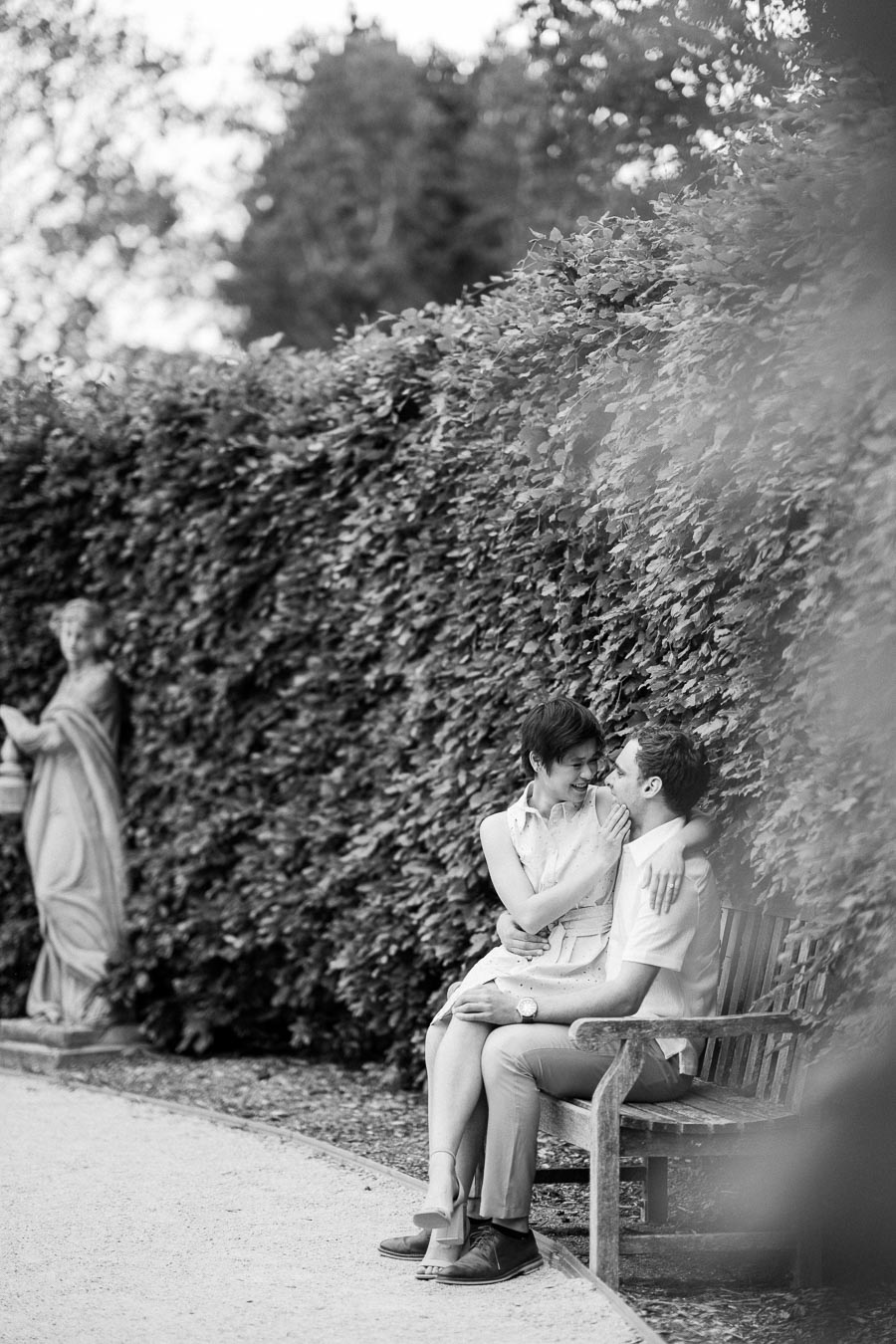 A romantic black and white photograph of a couple sitting closely on a wooden bench in a garden, surrounded by lush greenery, with a classical statue in the background, capturing a moment of joy and affection.