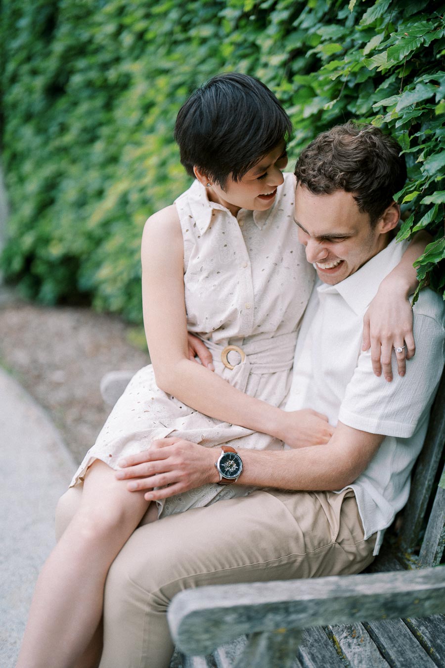 A couple sitting on a bench surrounded by lush green ivy, sharing a joyful and intimate moment together. The woman wears a sleeveless light dress while the man is dressed in a white shirt, both smiling and embracing one another.