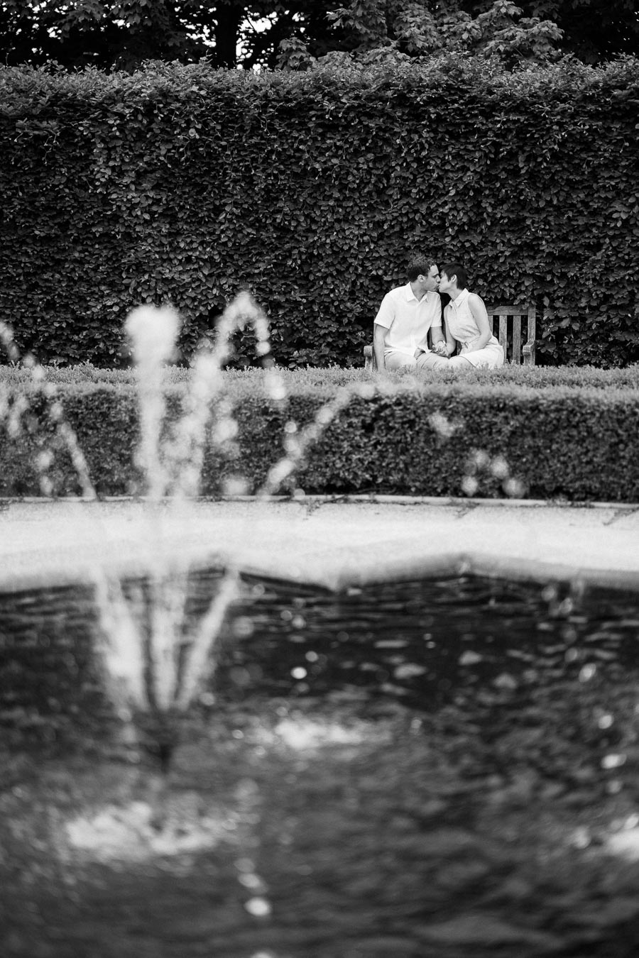 Black and white photo of a couple kissing on a bench by a hedge, with a fountain in the foreground.