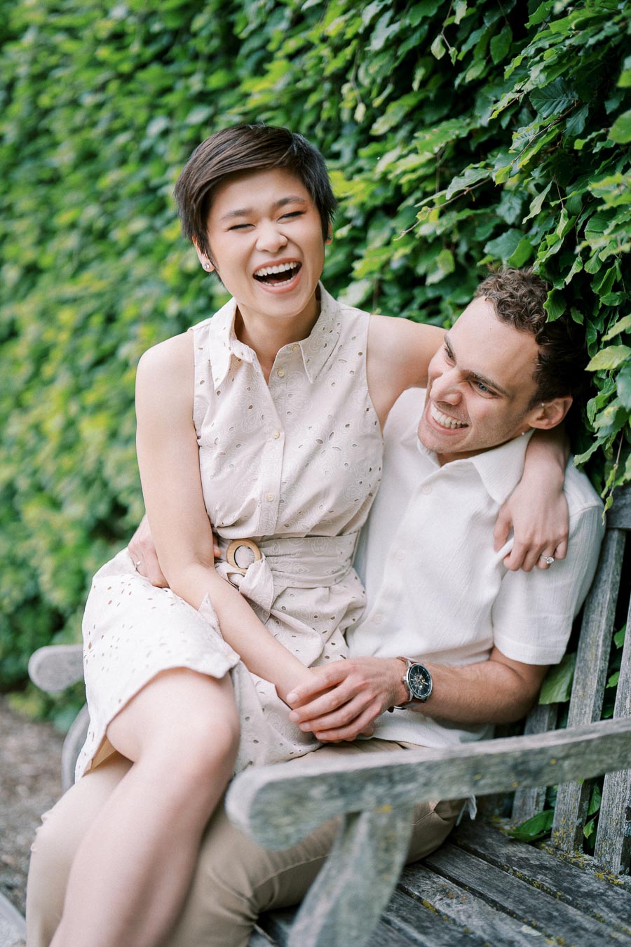A joyful couple laughing together on a wooden bench, set against a backdrop of lush green foliage.