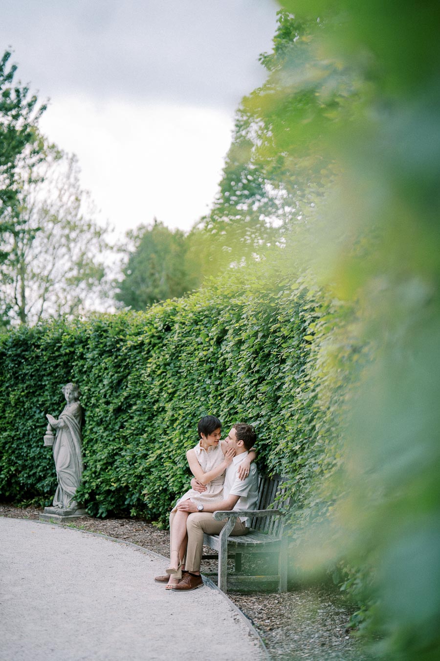 A couple sitting lovingly on a bench in a lush green garden with a statue in the background.