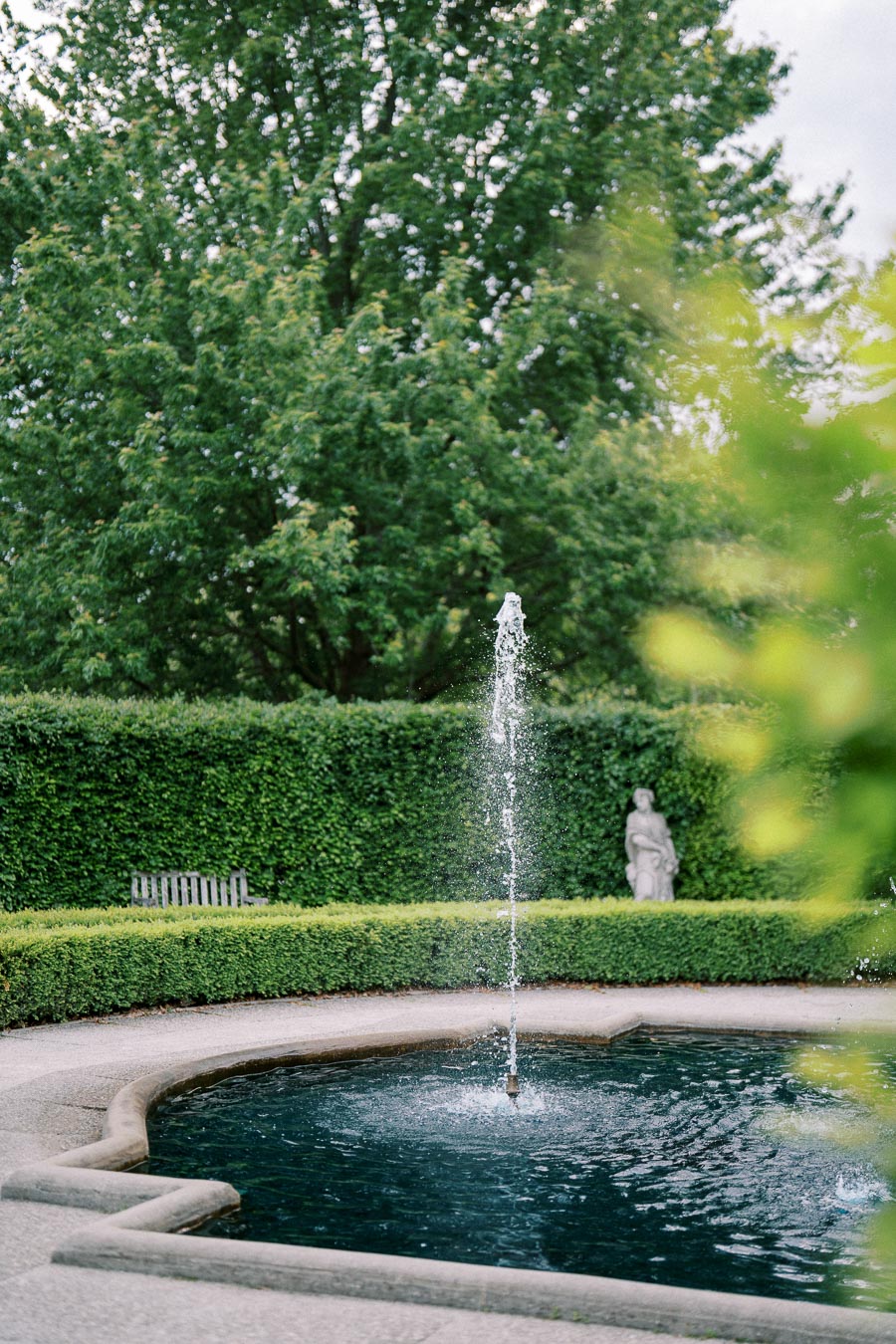 Elegant garden fountain surrounded by lush greenery and a statue in the background, creating a serene outdoor oasis.