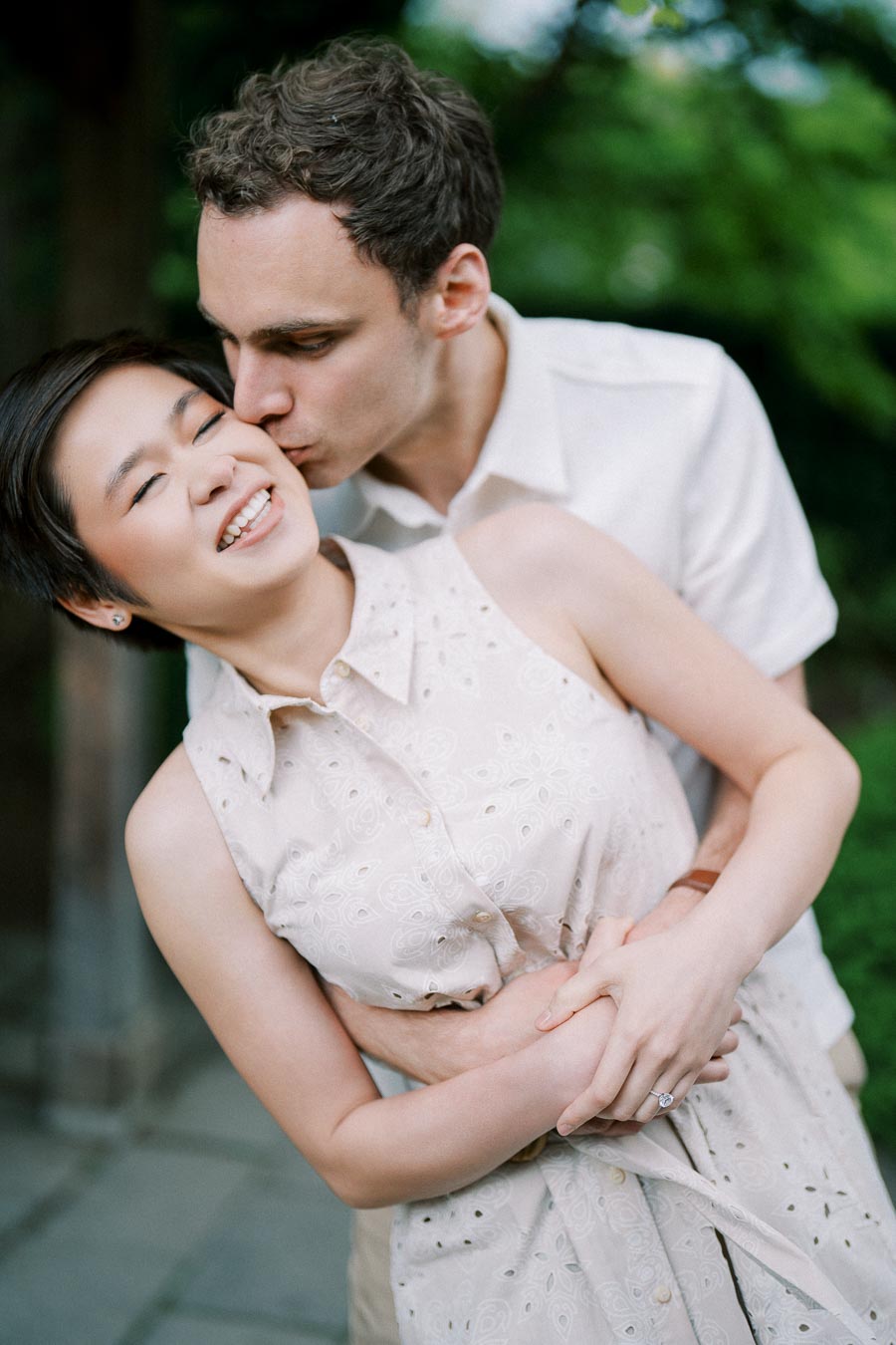 Happy couple embraces outdoors, with man kissing woman's cheek and woman smiling joyfully, showcasing love and affection in a garden setting.