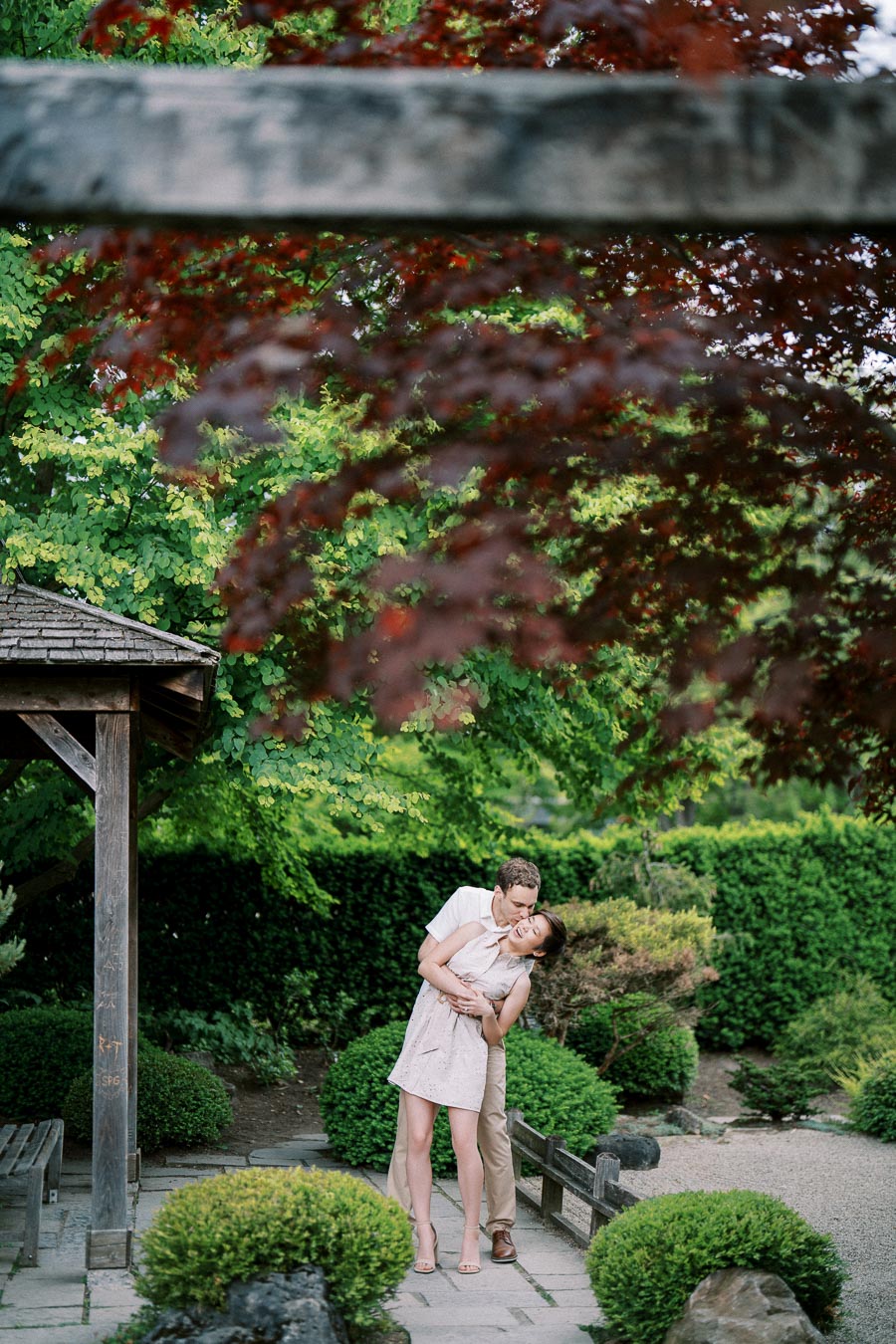 A couple embracing in a lush garden setting, surrounded by vibrant greenery and red leaves, under a wooden pergola.
