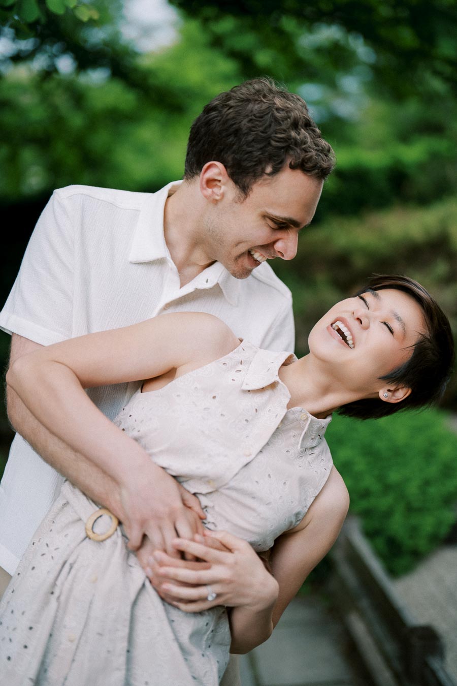 Couple laughing and embracing outdoors, surrounded by greenery, wearing light-colored clothing, enjoying a joyful moment in a natural setting.