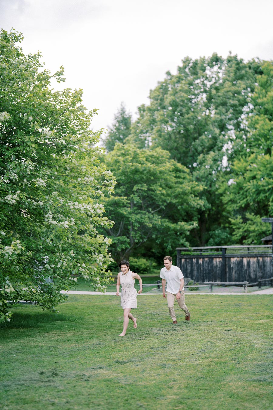 A man and woman running joyfully in a green park surrounded by lush trees, enjoying a sunny day outdoors.