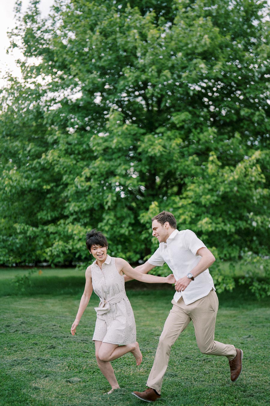 A happy couple enjoying a playful run on a lush green lawn in a park, surrounded by vibrant trees on a sunny day.