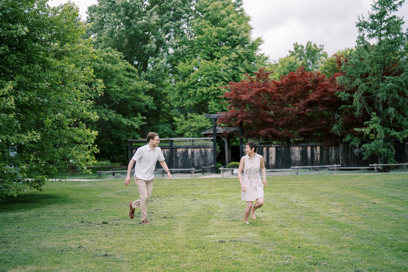 A couple joyfully running on green grass in a lush park, surrounded by vibrant trees and a wooden structure in the background.
