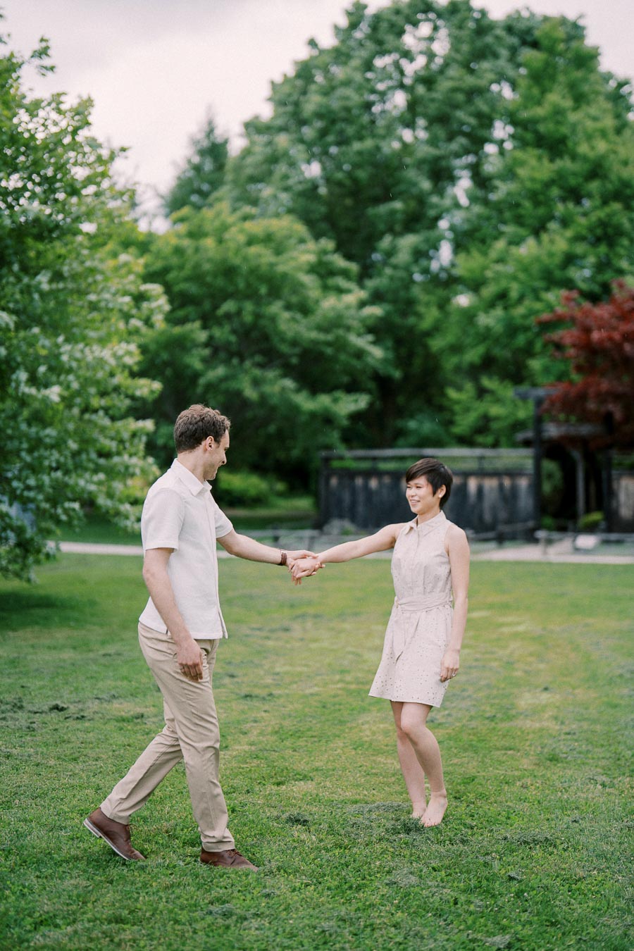 A couple holding hands and walking on a grassy field with lush green trees in the background.