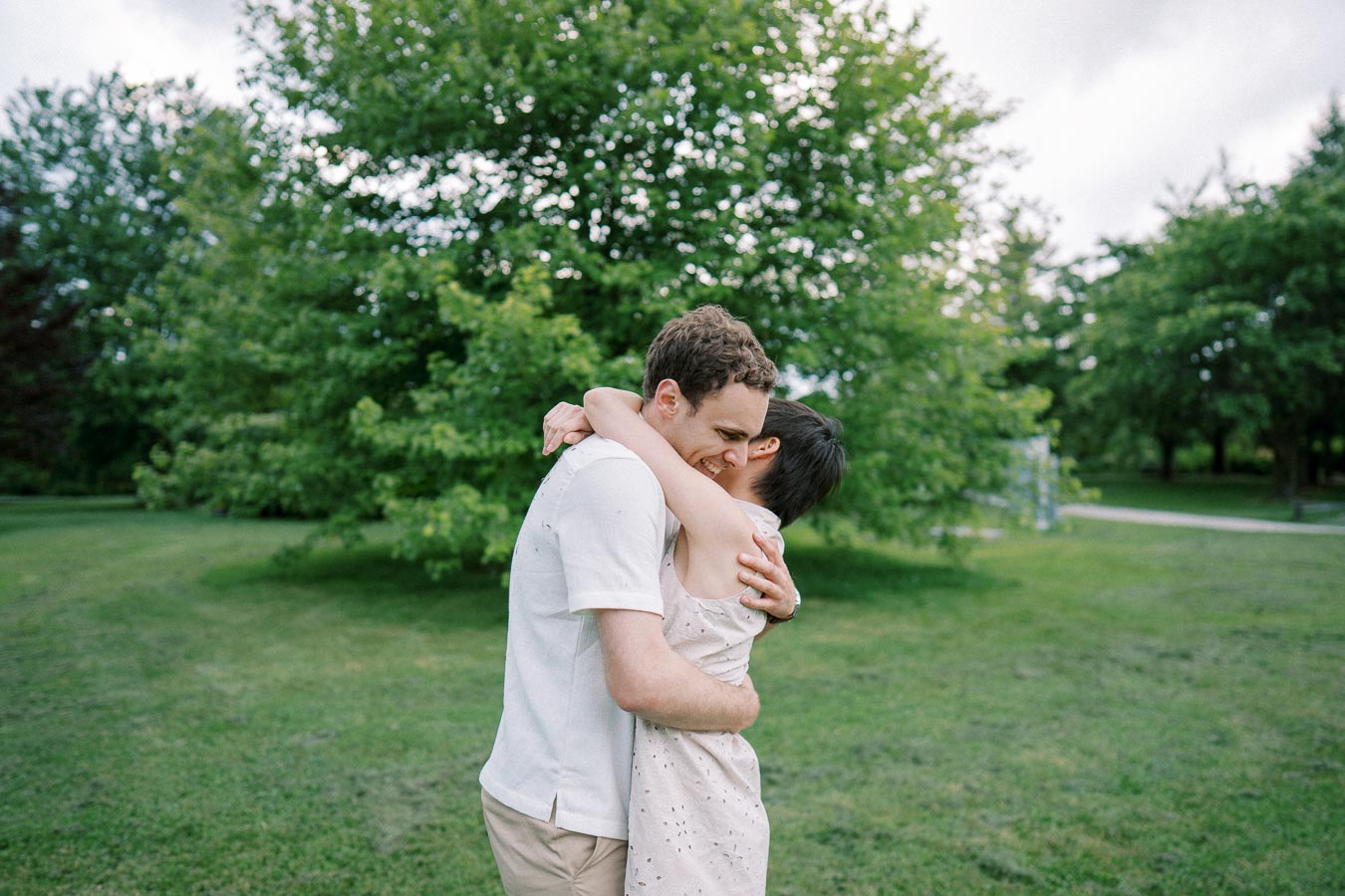 A couple embracing affectionately outdoors in a lush green park, surrounded by vibrant trees and open grass under a cloudy sky.