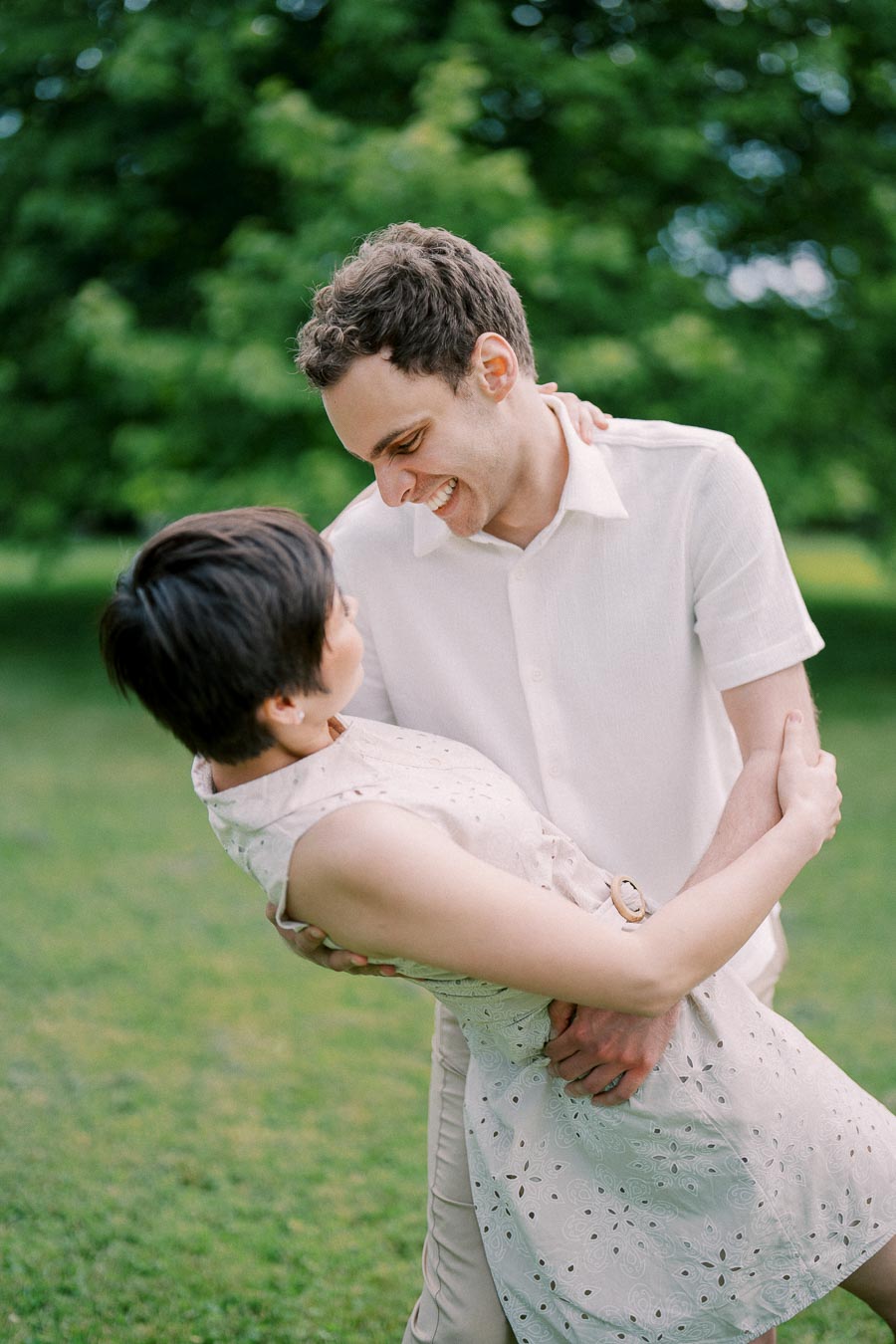 A joyful couple dancing in a green park, the man in a white shirt playfully dips the woman in a light-colored dress, capturing a moment of happiness and romance.