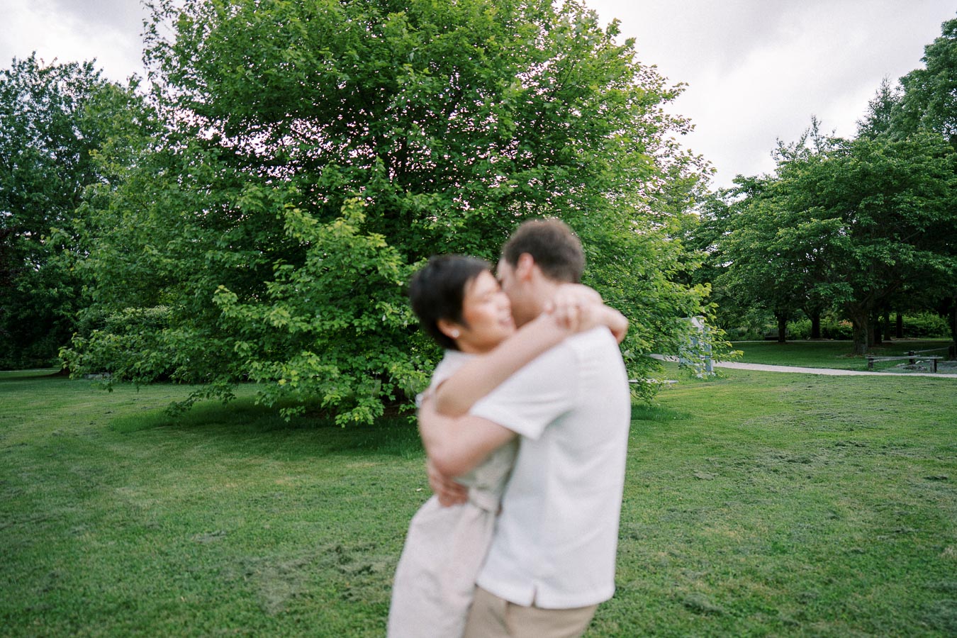 A couple embracing in a lush green park, surrounded by trees on a cloudy day.