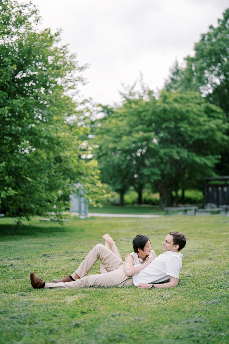 A couple relaxing on a grassy field, with lush green trees in the background, enjoying a peaceful day outdoors.