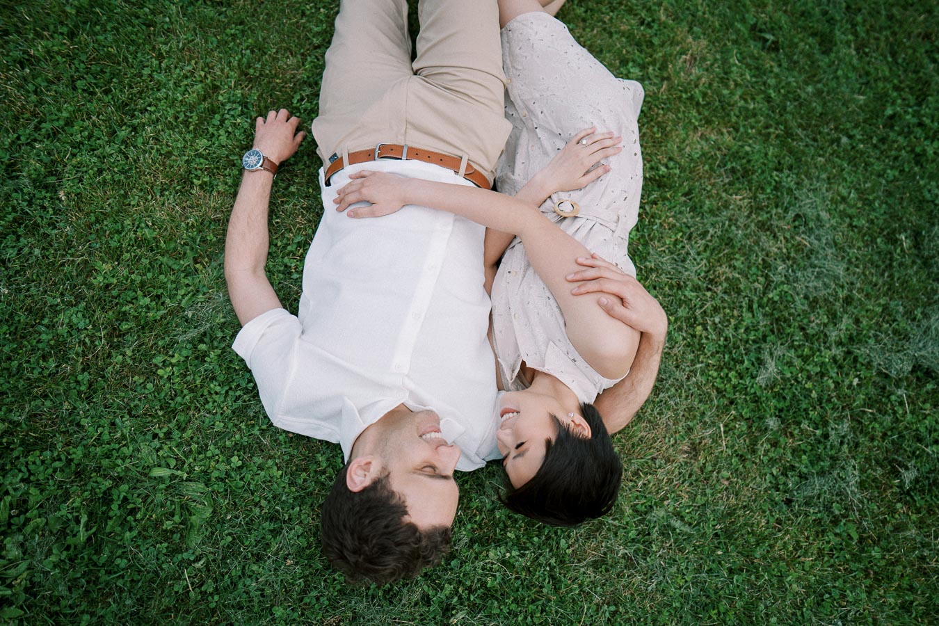 A happy couple lying on green grass, gazing into each other's eyes with smiles, showcasing love and togetherness in a serene outdoor setting.