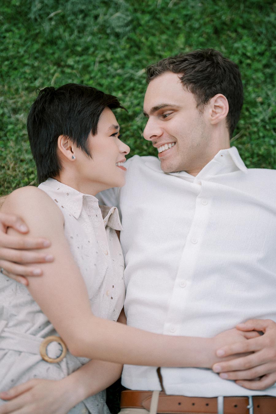 A happy couple lying on grass, smiling at each other, wearing white outfits, embracing affectionately in a serene outdoor setting.