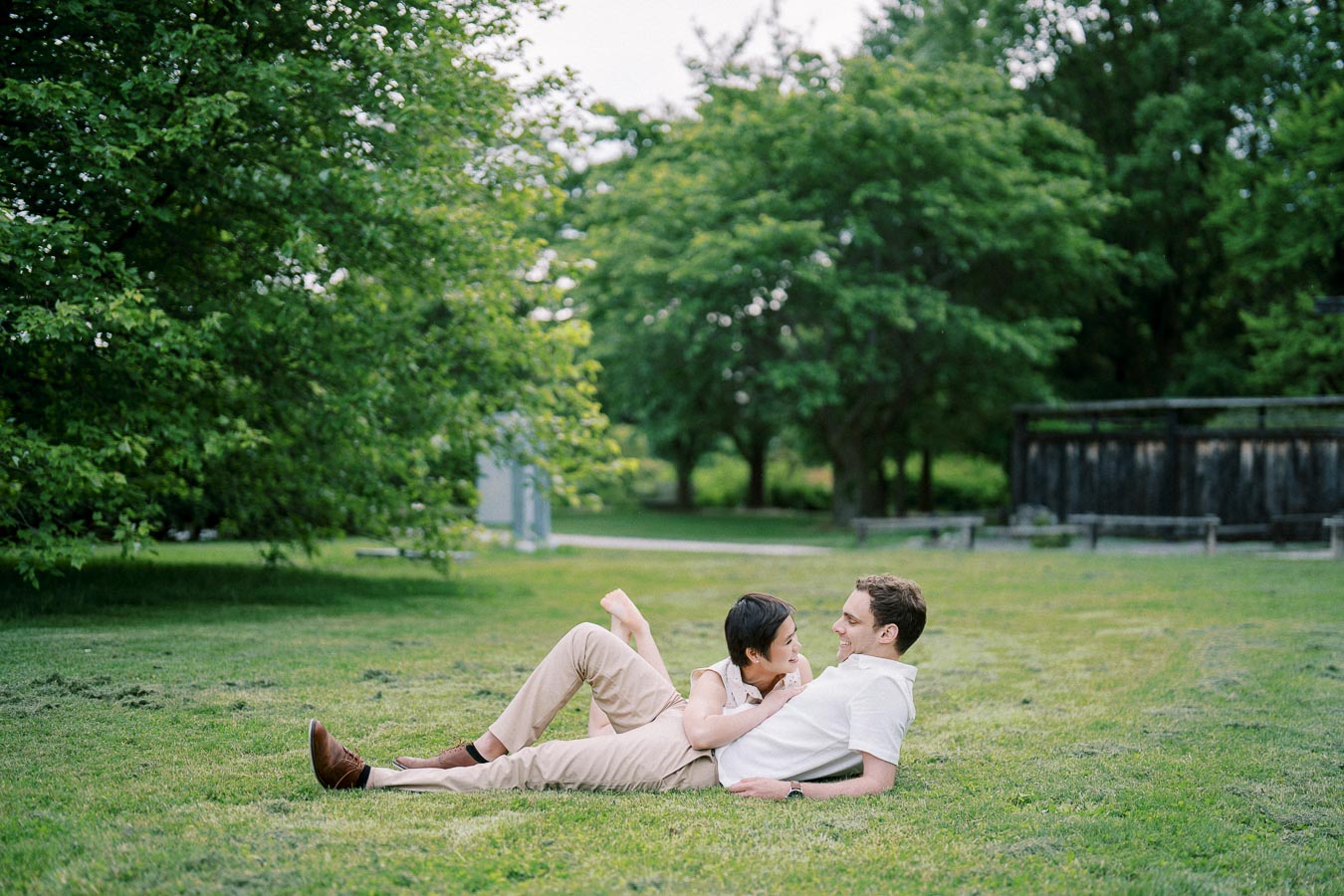 A couple relaxing on a grassy field in a park, surrounded by lush green trees, enjoying a peaceful moment together in nature.