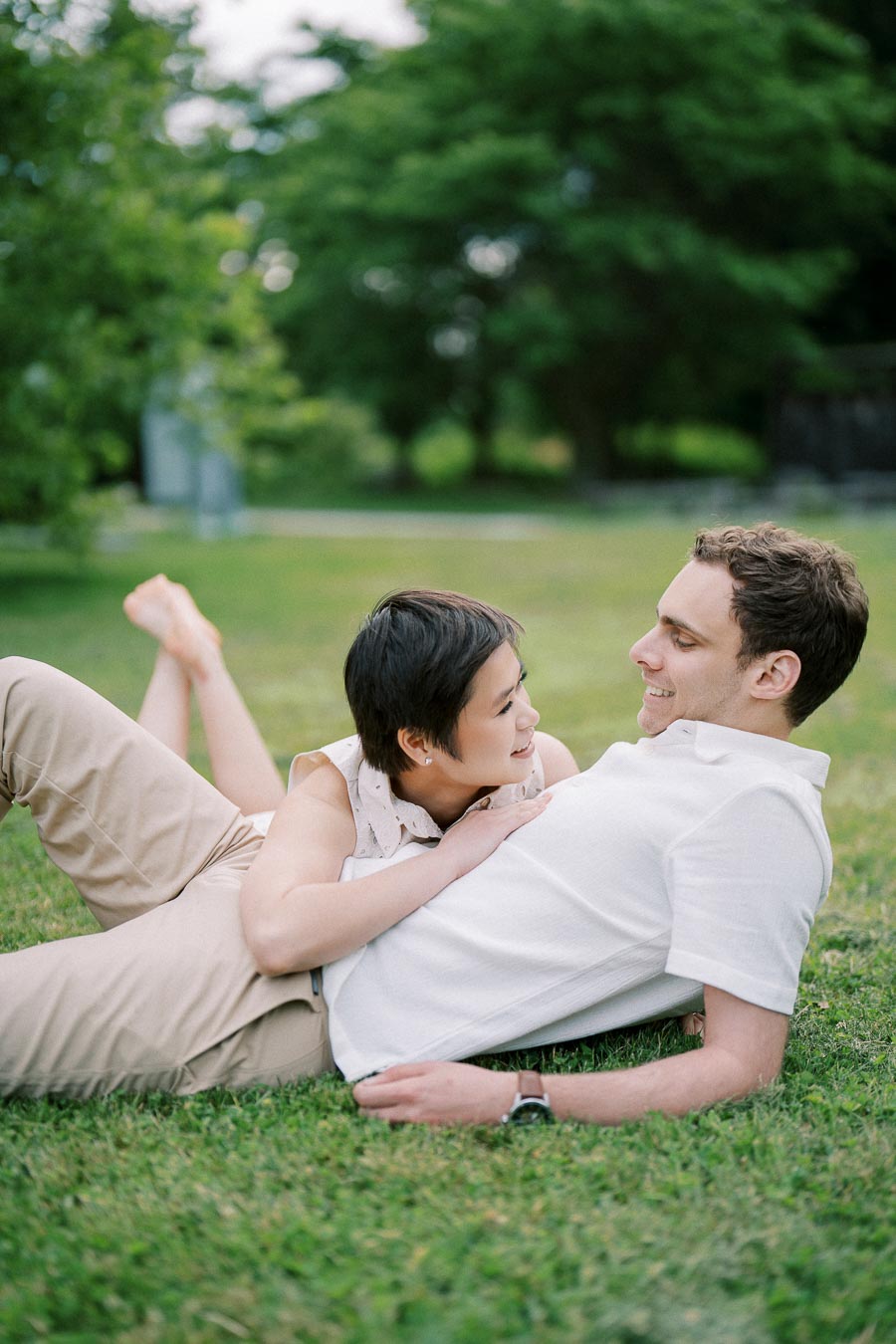 A happy couple lying on the grass in a park on a sunny day, smiling at each other, surrounded by lush greenery, showcasing a romantic outdoor moment.