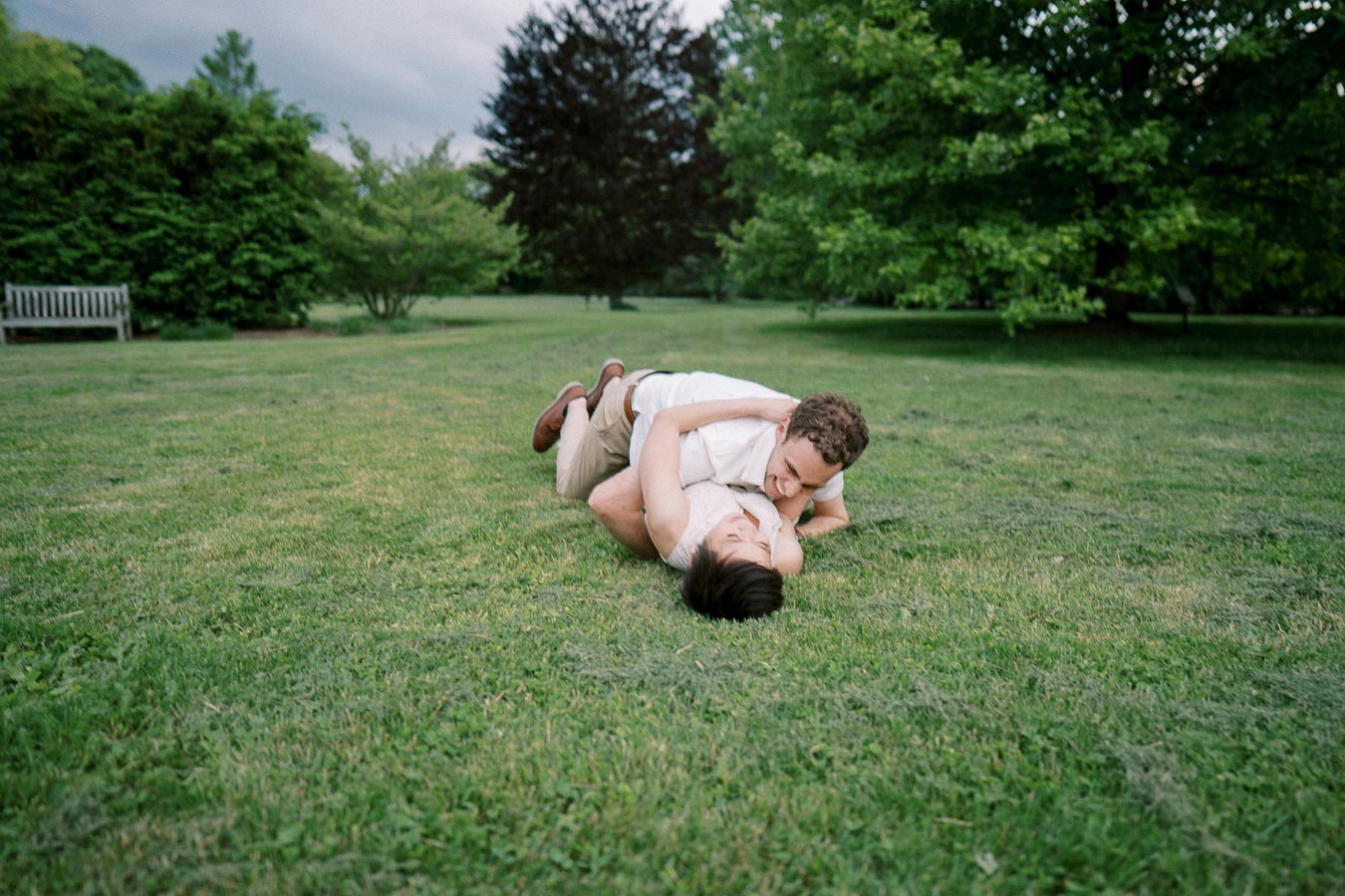 A couple lying on the grass in a park, embracing and smiling at each other, surrounded by lush greenery.